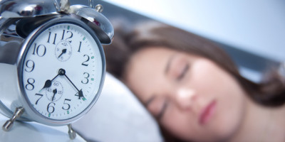 Unpünktlichkeit: Wecker steht auf 7.30 Uhr, im Hintergrund schläft eine Frau im Bett
