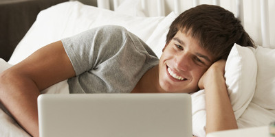 Illegale Downloads: Junger Mann liegt seitlich auf dem Bett und schaut auf seinem Laptop einen Film