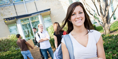 Junge Studentin steht vor dem Studentenwohnheim, im Hintergrund eine Gruppe von Studenten