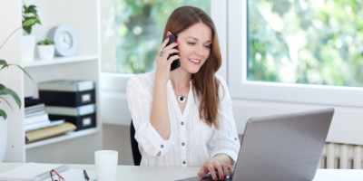 Überstunden bei Teilzeit: Ein Frau sitzt im Büro vor einem Laptop und telefoniert mit einem Smartphone.