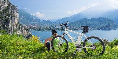 E-Bike nachrüsten: Diese Regelungen sind zu beachten. Eine Frau sitzt in einer Berglandschaft mit Ihrem E-Bike auf einer mit Gras bewachsenen Klippe und blickt einen türkisen See herunter.