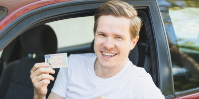 Führerschein auf Probe: Besondere Regeln für Fahranfänger. EIn junger Mann guckt lächelnd aus einem Auto und hält einen Führerschein in seiner Hand.