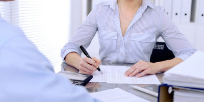 Zweitwohnsitz anmelden: Darauf müssen Sie achten. Eine Frau mit gestreifter Bluse sitzt an einem Bürotisch und zeichnet auf einem Dokument.