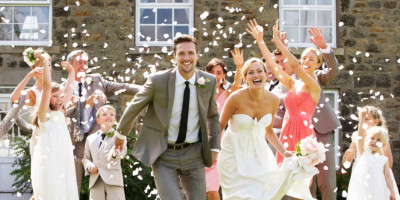 Braucht man Trauzeugen zum Heiraten? Ein frisch vermähltes Ehepaar freut sich. Im Hintergrund sieht man die Hochzeitsgesellschaft, die Blütenblätter wirft.