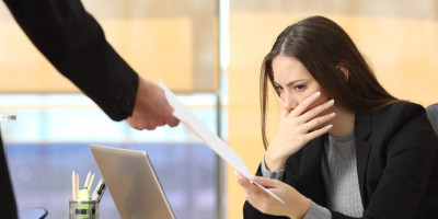Kündigung formulieren: Darauf müssen Arbeitgeber achten. Eine Frau im Blazer sitzt am Arbeitsplatz und hält sich erschrocken die Hand vor den Mund während ihr eine andere Person ein Dokument überreicht.