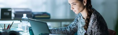 Überstunden: Frau sitzt vor ihrem Laptop, alleine in dunklem Büro