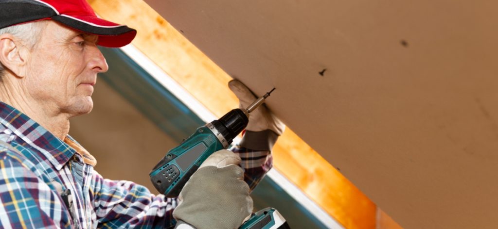 Drywall construction, attic renovation. Man fixing drywall suspended ceiling to metal frame using electrical screwdriver