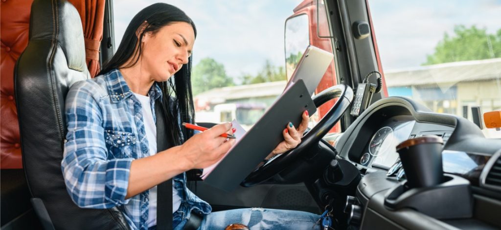 Eine junge Frau sitzt auf dem Fahrersitz eines Lkw und hält einen Stift und Mappen in der Hand